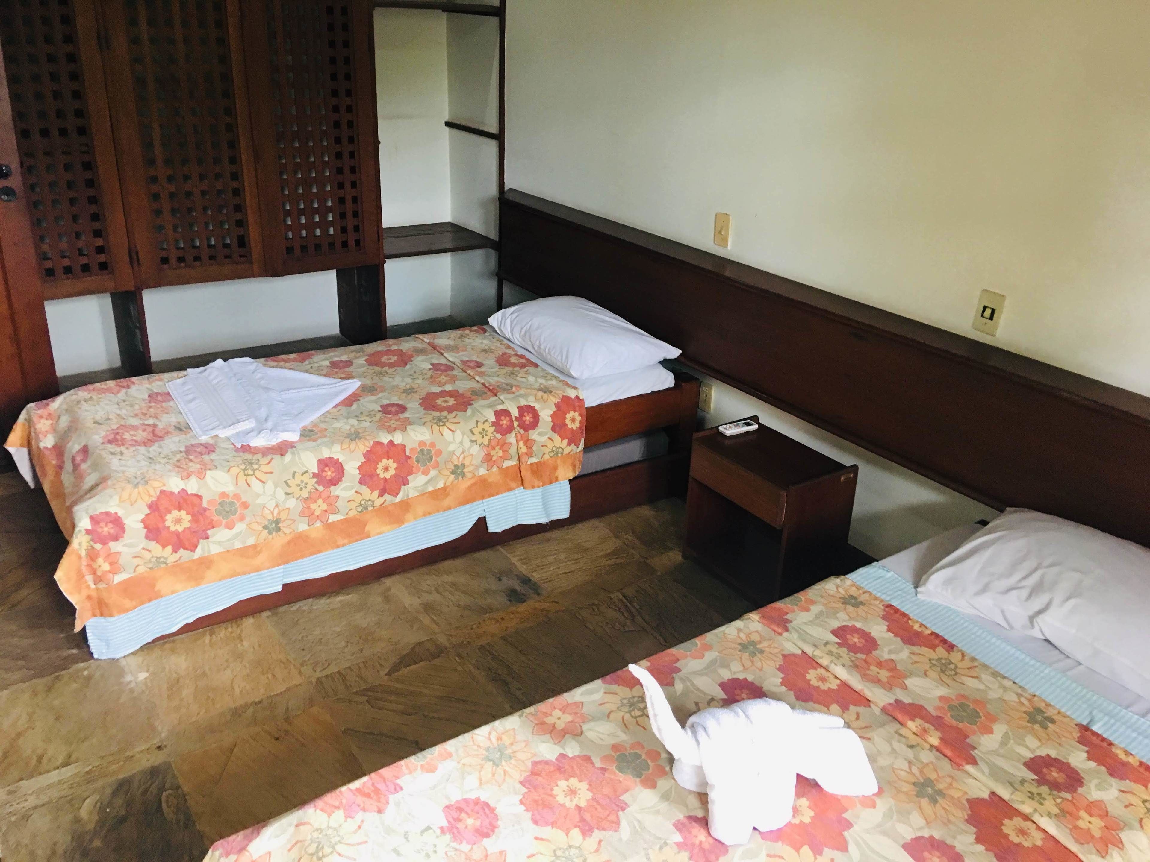 A hotel room with two beds, wooden headboards, a small bedside table with a remote control, and a wooden floor.