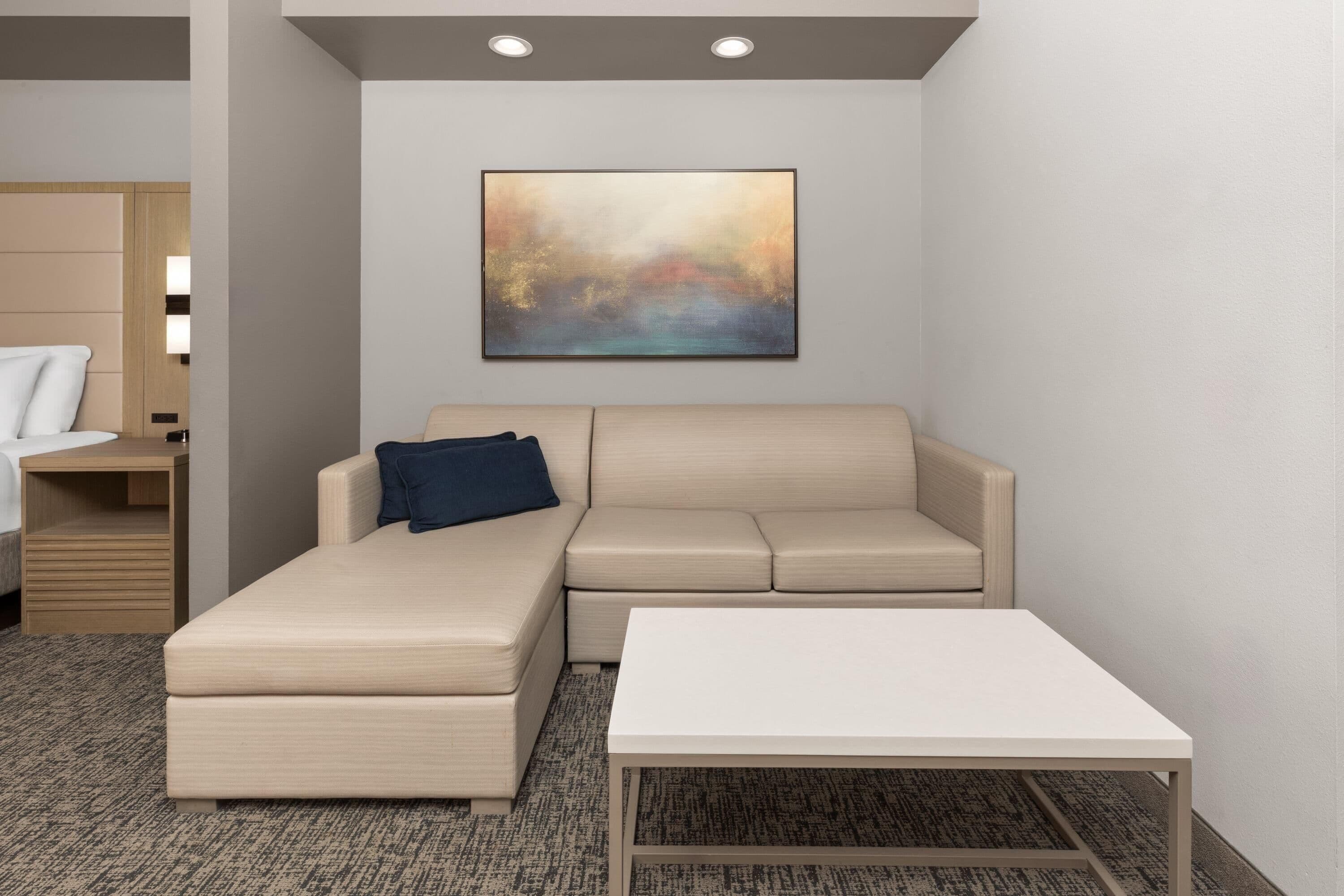 A modern hotel room with a beige sectional sofa, a white coffee table, and a large abstract painting on the wall.