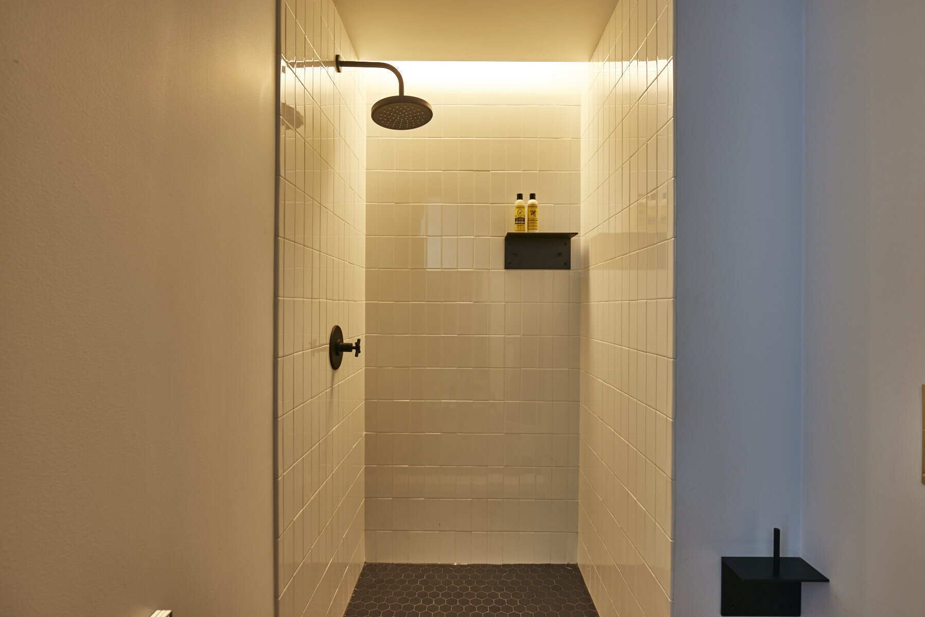 A narrow, tiled shower area with a handheld showerhead, a small shelf with toiletries, and a door leading into the space.