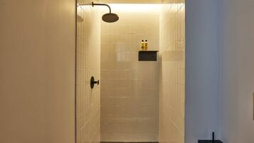 A narrow, tiled shower area with a handheld showerhead, a small shelf with toiletries, and a door leading into the space.