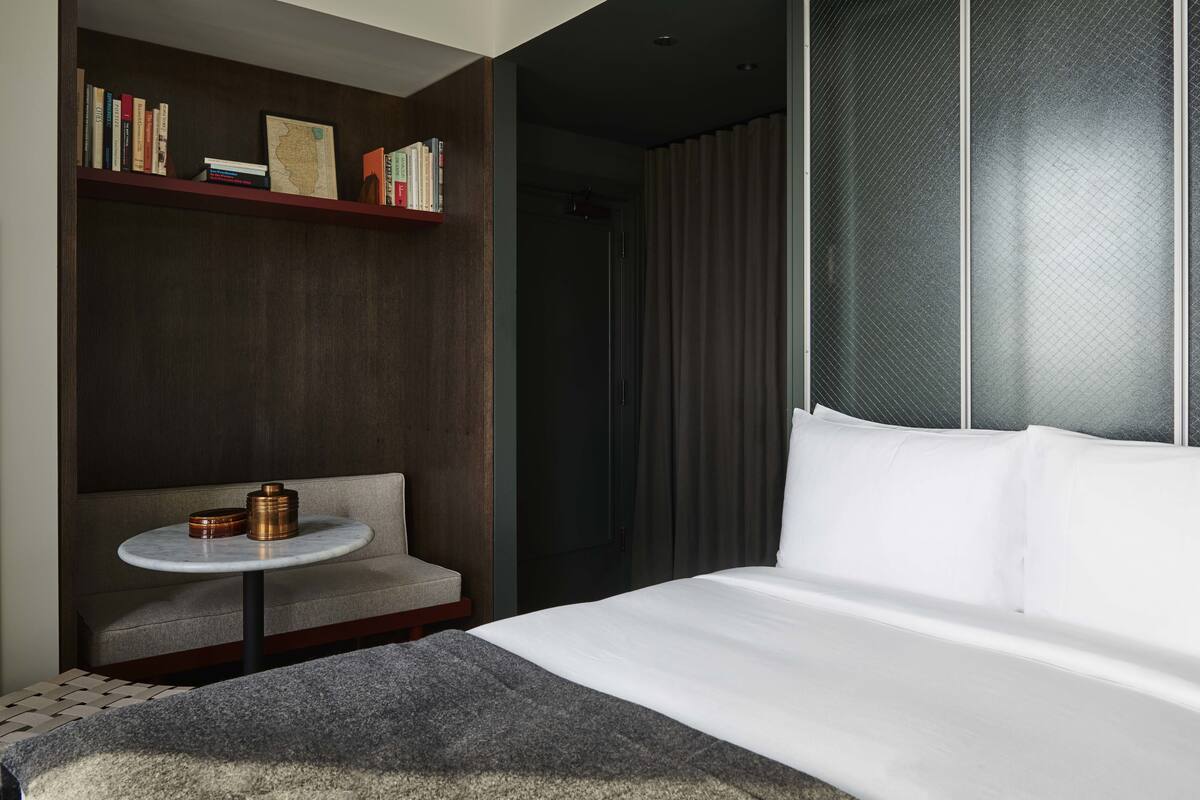 A hotel room with a bed, a small round table with a lamp, a bookshelf with books, and a large window with curtains.