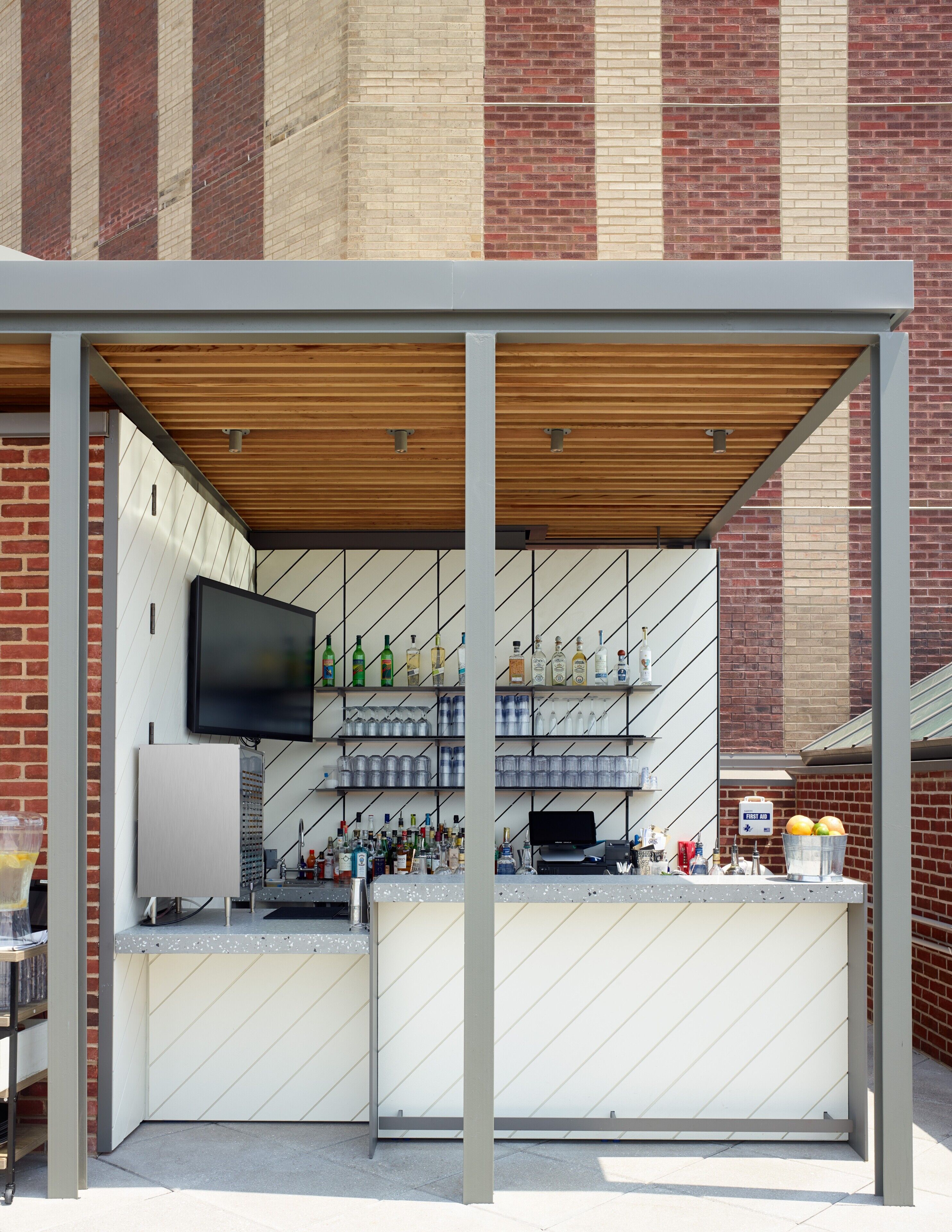 Zona de bar al aire libre con techo de madera, estantes con botellas, un televisor y un letrero que indica primeros auxilios.