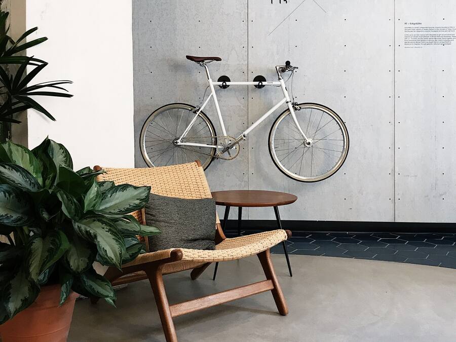 A room with a white wall, a mounted bicycle, a wicker chair, a small round table, and a potted plant.