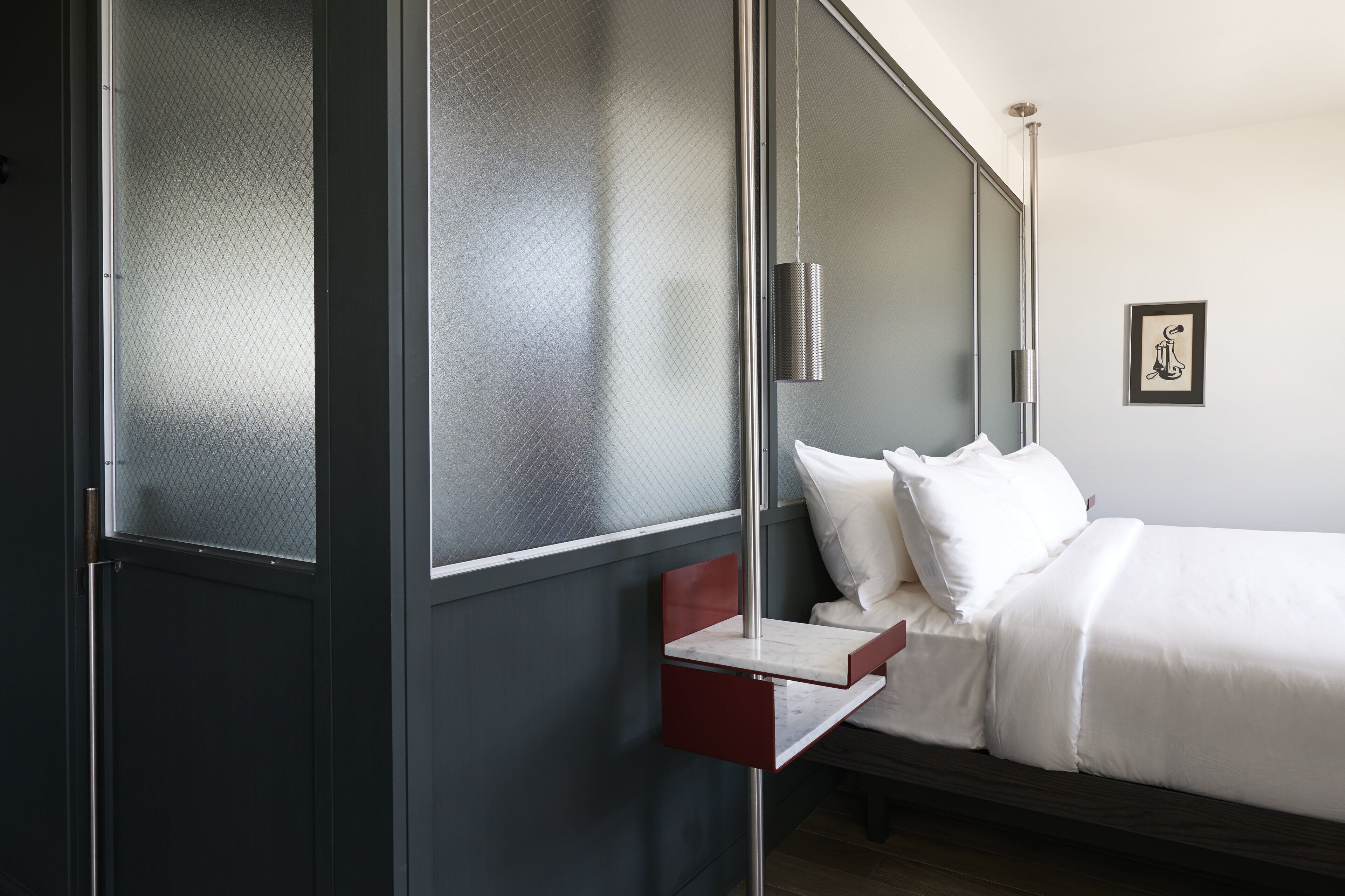 A modern hotel room with a bed, a small bedside table, a nightstand, and a wall-mounted lamp.