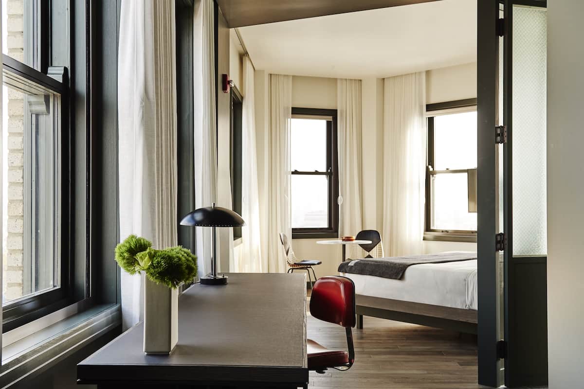 A hotel room with a bed, a desk with a lamp, a chair, and a small table.