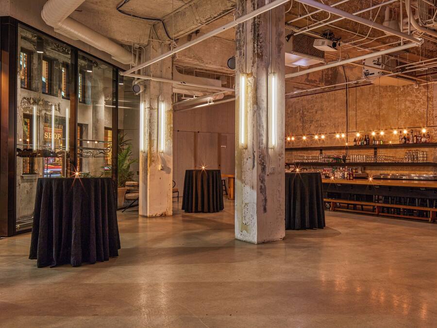 Zona de bar de estilo industrial con techos expuestos y pilares de hormigón, con una barra alargada y taburetes.