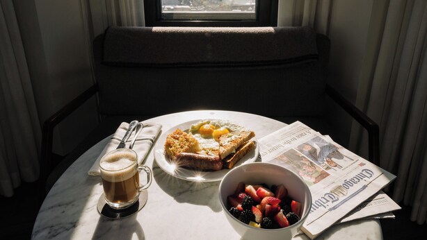A breakfast setting with a plate of toast, a bowl of fruit, a cup of coffee, and a newspaper on a table by a window with a city view.