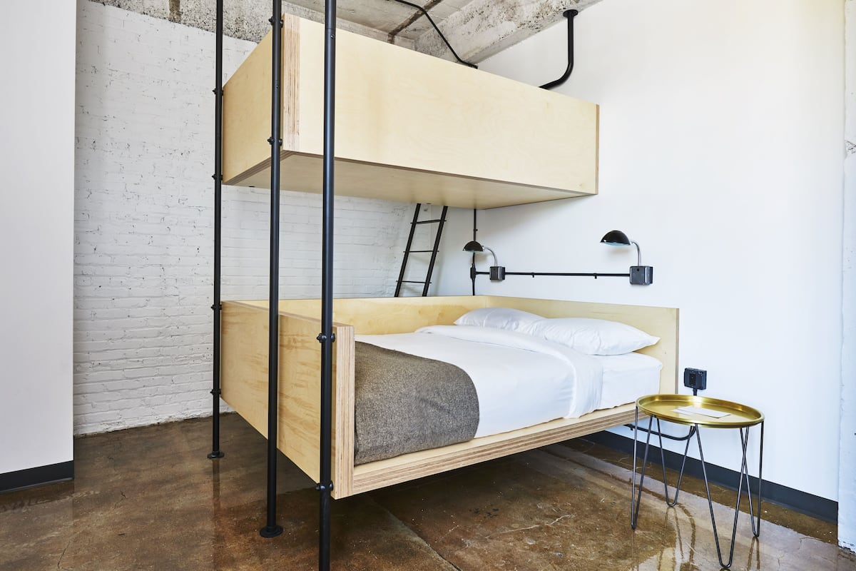 A bunk bed with a ladder, a bedside table, and a wall-mounted lamp in a room with exposed brick walls and a concrete floor.