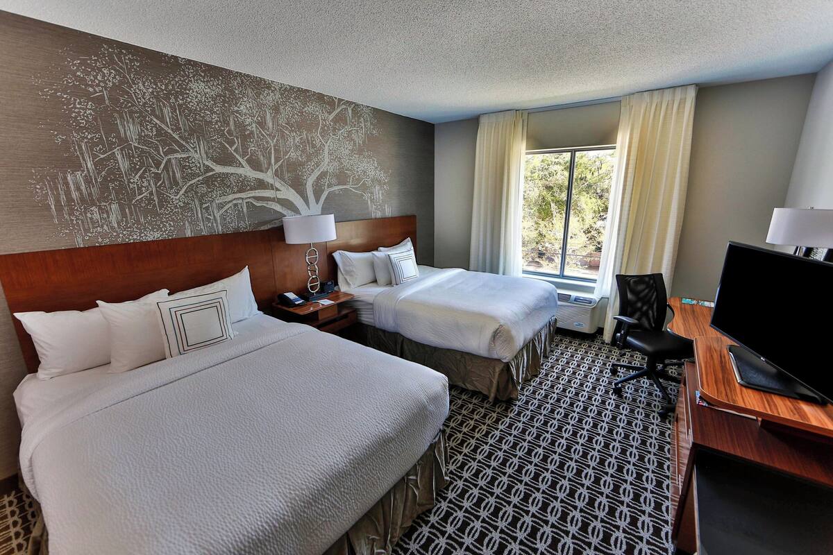 A hotel room with two beds, a desk with a TV, a large window with curtains, and a wall mural.