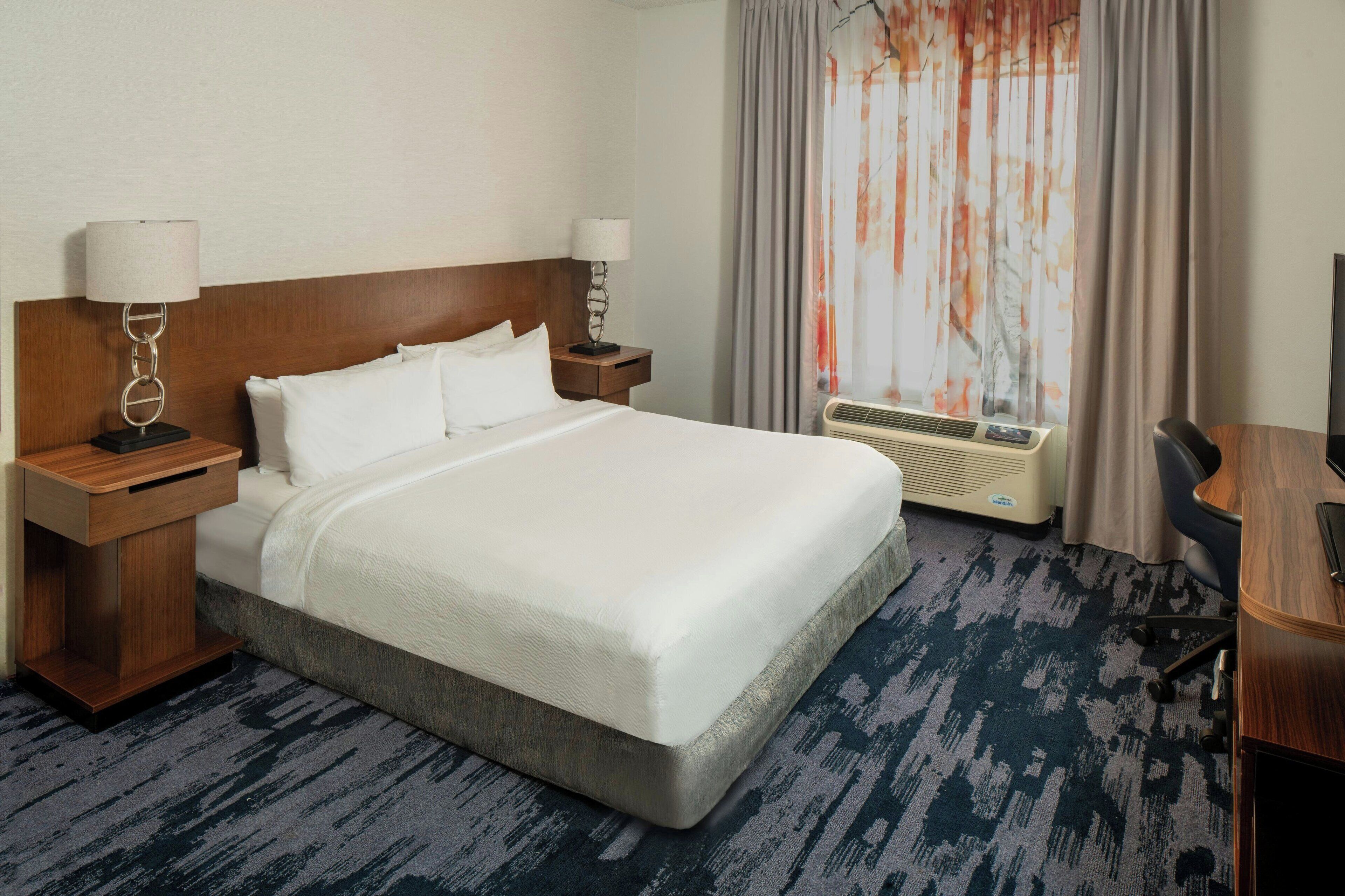 A hotel room with a large bed, two bedside tables with lamps, a desk, an air conditioning unit, and a window with curtains.