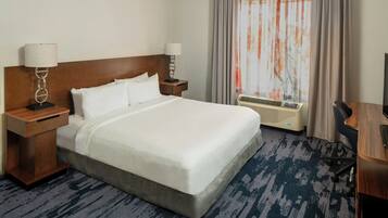 A hotel room with a large bed, two bedside tables with lamps, a desk, an air conditioning unit, and a window with curtains.
