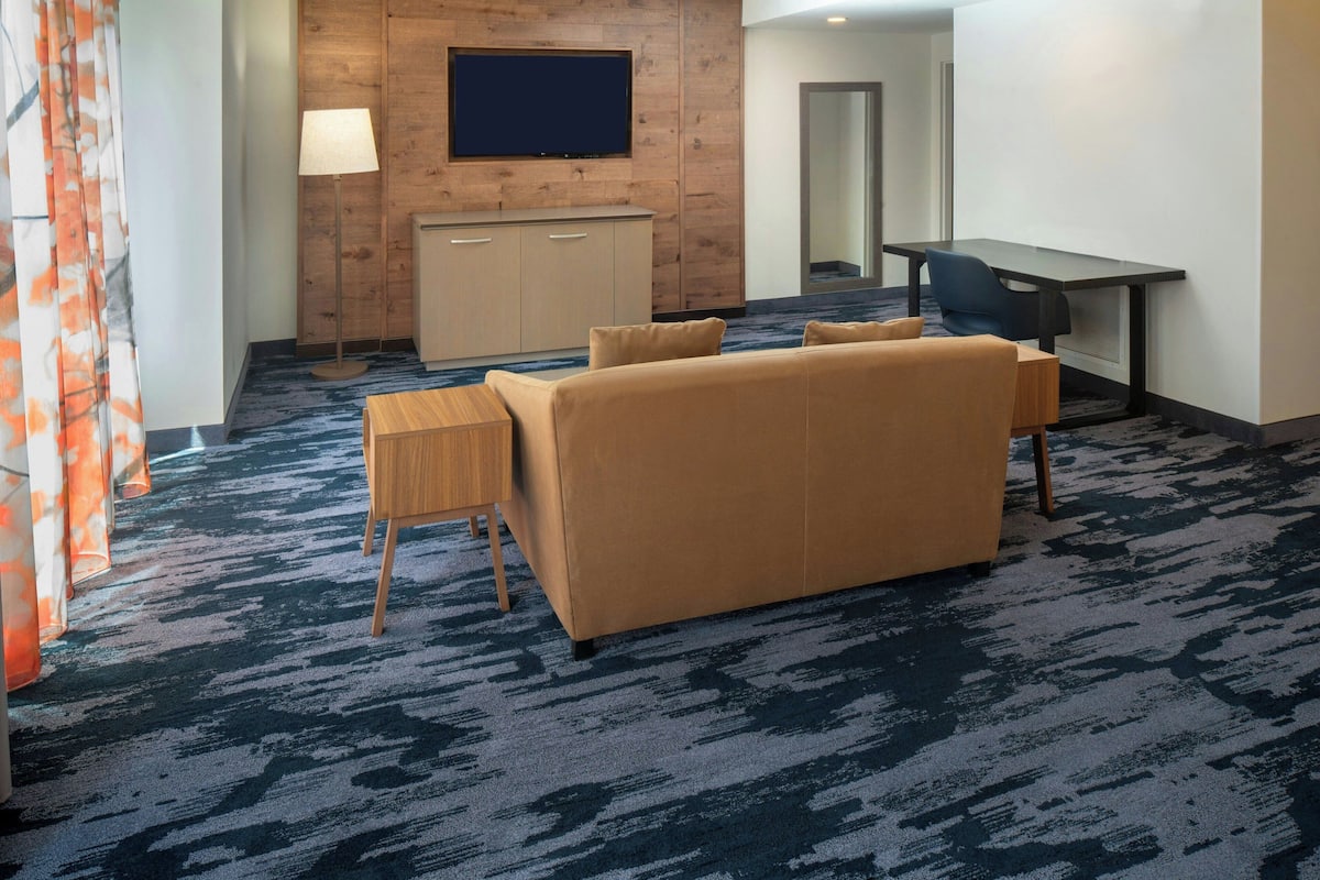 A hotel room with a sofa, a small wooden table, a desk, a television mounted on a wooden panel wall, and a carpeted floor.