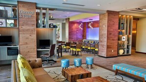 Eine Hotel-Lobby mit einer Couch, einem Couchtisch und einer Sitzecke mit Sesseln und Hocker. Es gibt eine Rezeption mit Computer und Fernseher.