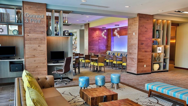Een hotel lobby met een bank, salontafel en zithoek met stoelen en poefs. Er is een receptiebalie met een computer en een televisie.