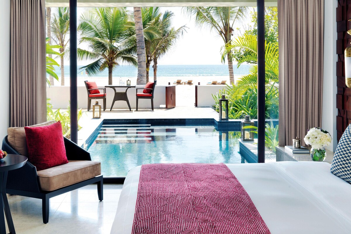 A hotel room with a bed, a small table, a chair, and a view of the ocean.