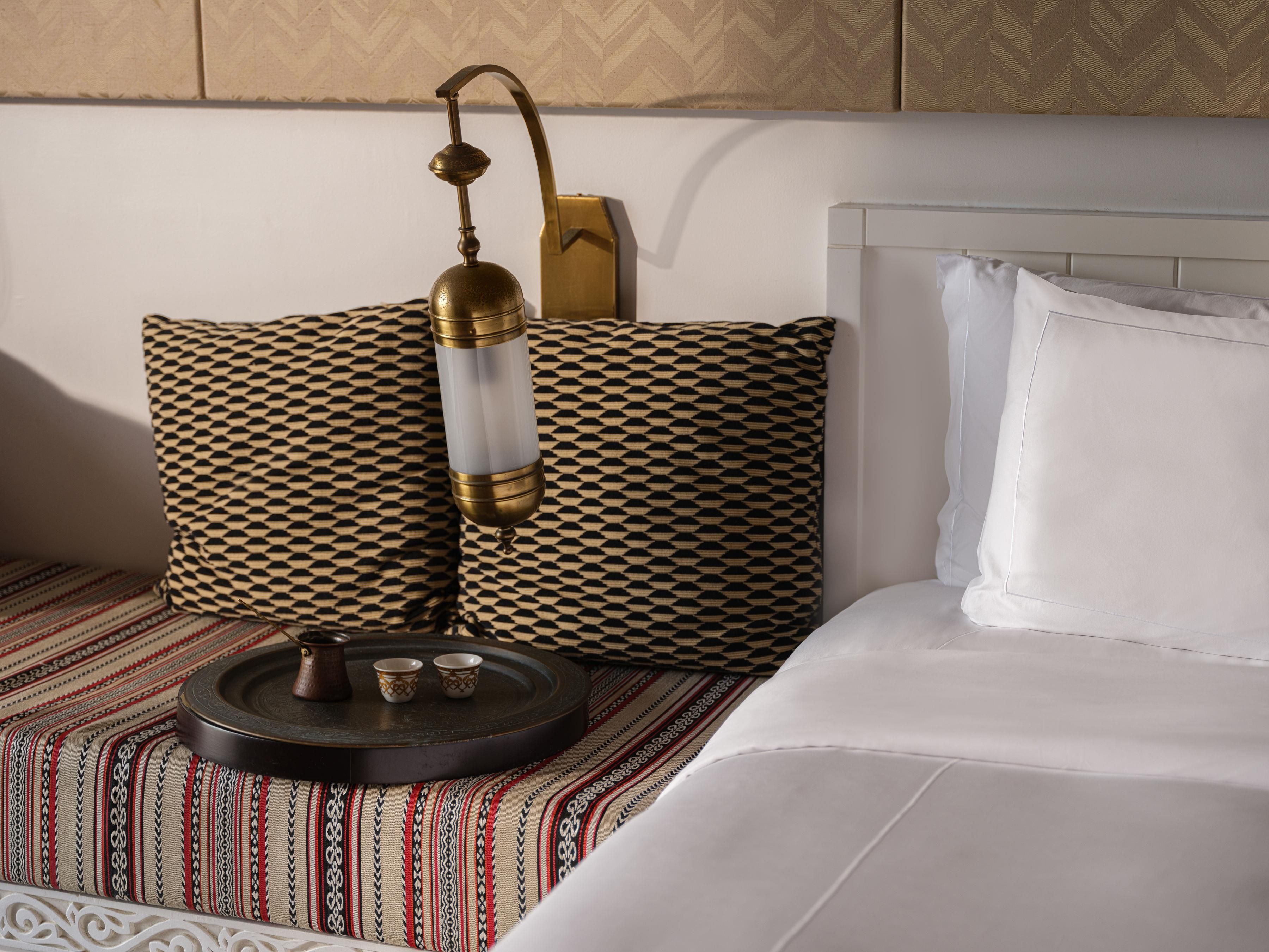 A hotel room with a bed, patterned pillows, a bedside table with a lamp, and a tray with cups.
