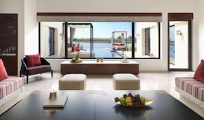 A modern living room with a large window overlooking a pool and a scenic view. There is a sofa, a coffee table with fruit, and a bookshelf. - Al Baleed Resort Salalah by Anantara (Salalah)