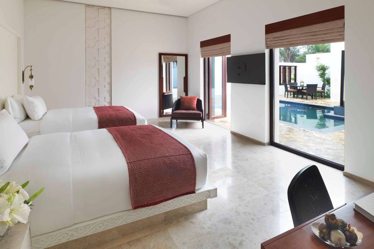A hotel room with two beds, a desk, a chair, a dining table, and a view of a swimming pool.