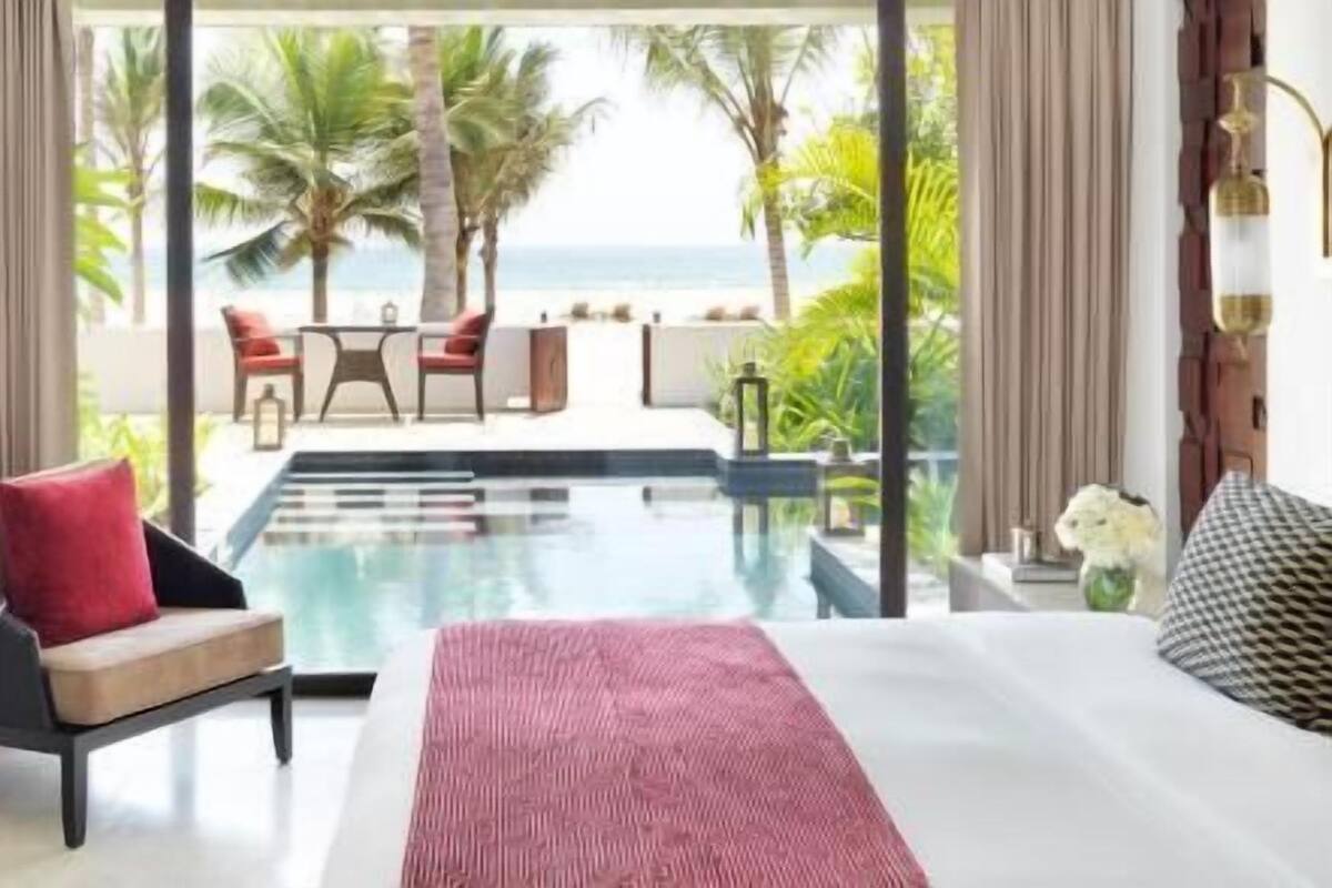 A hotel room with a large bed, a small table, a chair, and a view of the ocean and palm trees.