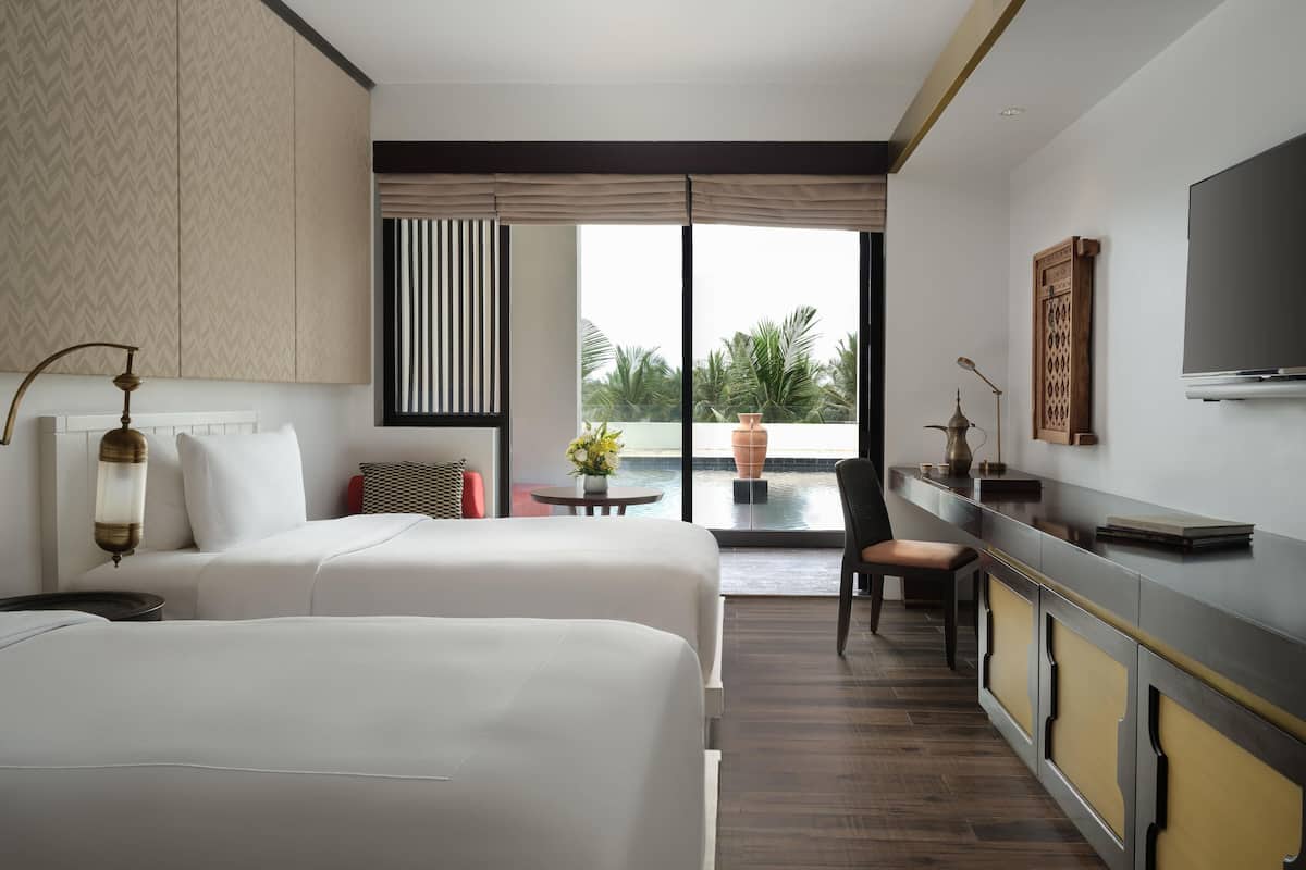 A hotel room with two beds, a desk, a chair, a TV, a window with a view of palm trees, and a potted plant.