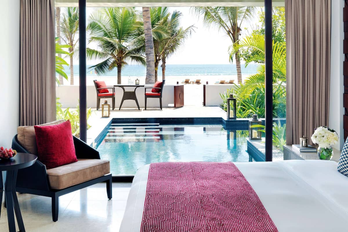 A hotel room with a large bed, a pool, and a view of the ocean and palm trees.