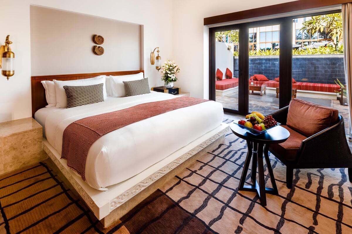 A hotel room with a large bed, a small table with fruit, a chair, and a view of the outdoor seating area through the glass doors.