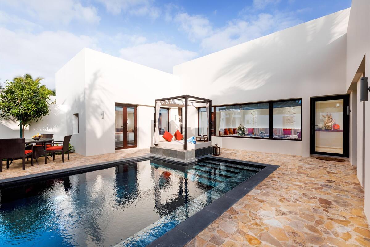 A modern outdoor pool area with a stone floor, a glass-enclosed seating area, and a dining setup with chairs and a table.
