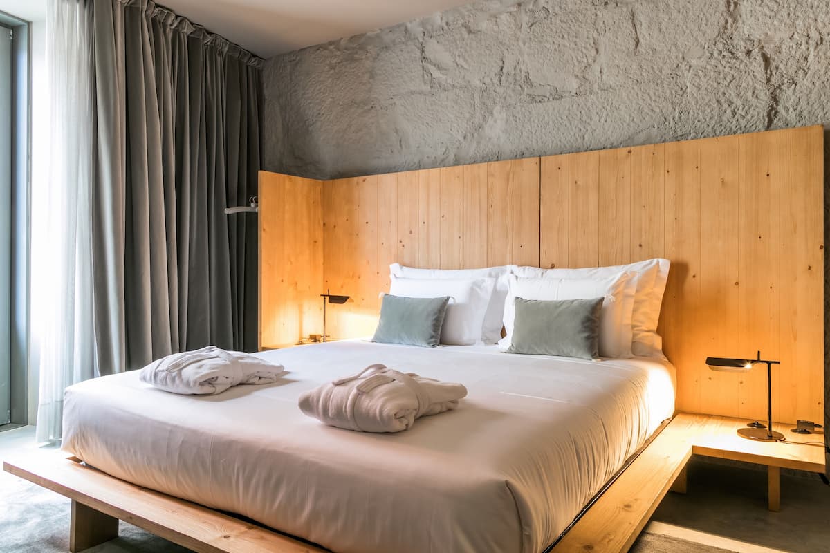 A modern bedroom with a wooden headboard, a large bed with white bedding, two bedside tables with lamps, and a view of the outdoors through a window with curtains.
