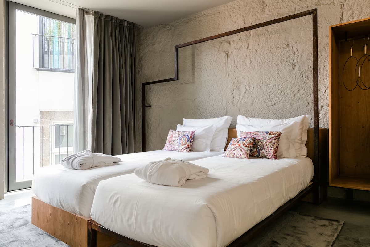 A double bed with white linens and colorful pillows, a wooden headboard, a large window with curtains, and a rustic textured wall.