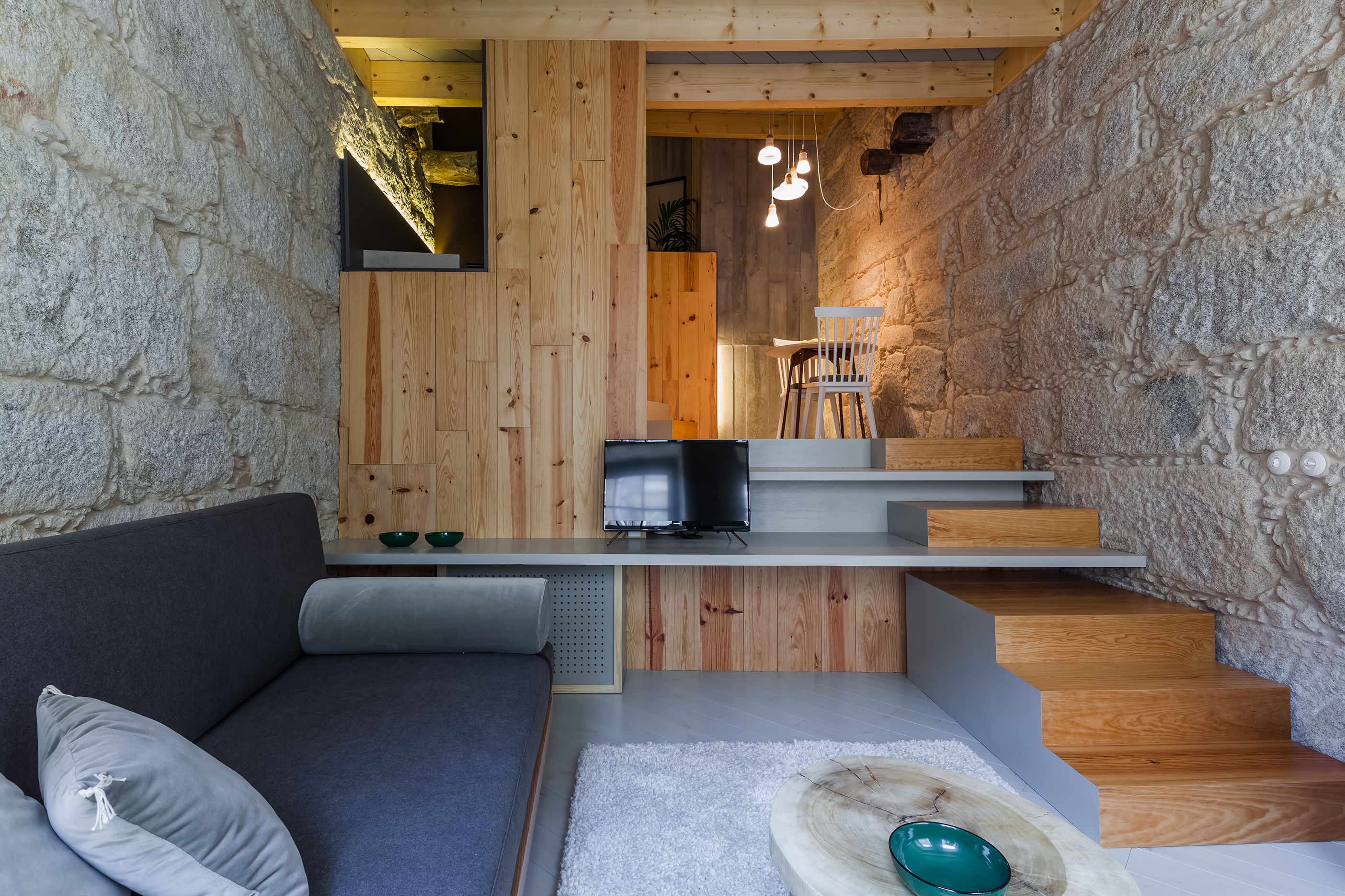 Un interior moderno con una pared de piedra, paneles de madera, un sofá gris, una pequeña mesa con un cuenco y una escalera.