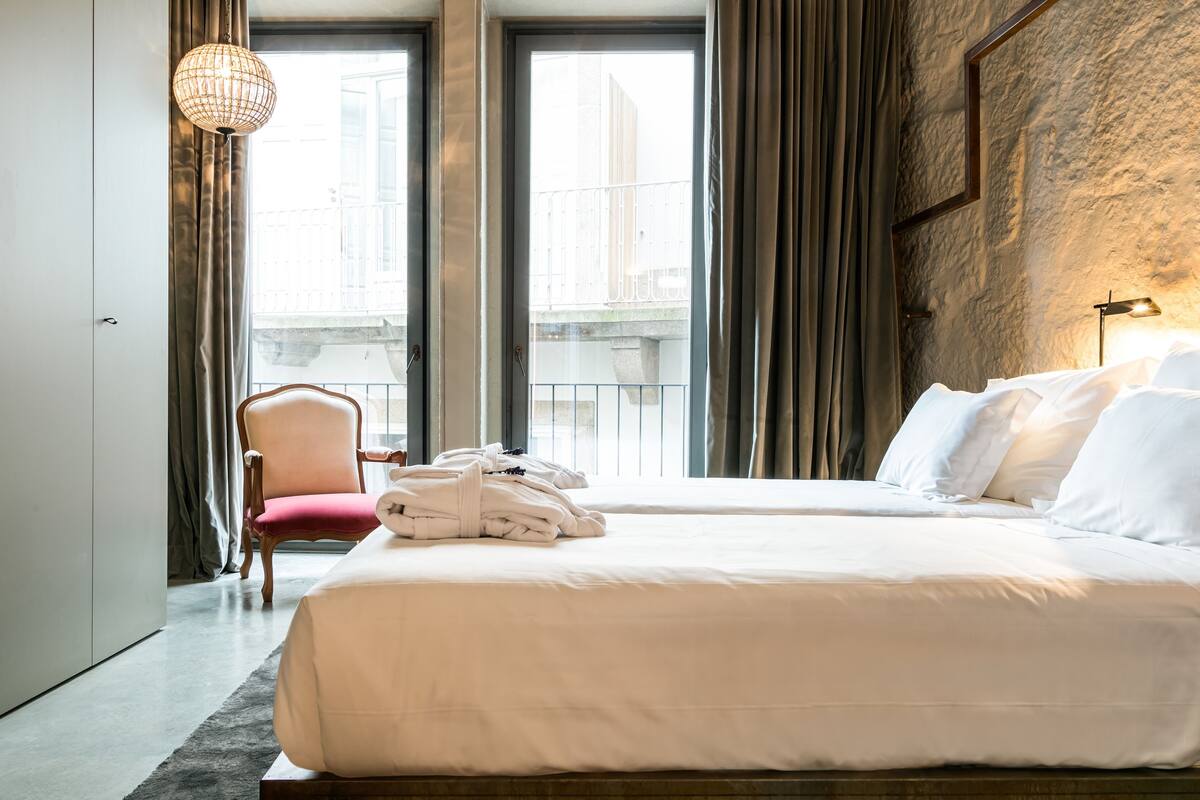 A hotel room with a large bed, a chair, a wardrobe, and a view of the city through large windows.