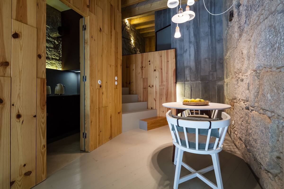A wooden interior with a round table and white chairs, a staircase, and a stone wall.