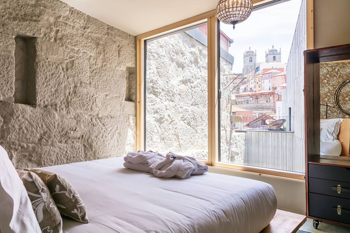 A bedroom with a stone wall, a large window with a view of buildings, a bed with white linens, and a wooden dresser.