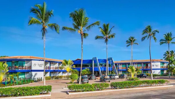 A hotel with a modern design, featuring a blue and white color scheme, multiple floors, and a parking area with several cars. - Sueds Plaza (Porto Seguro)