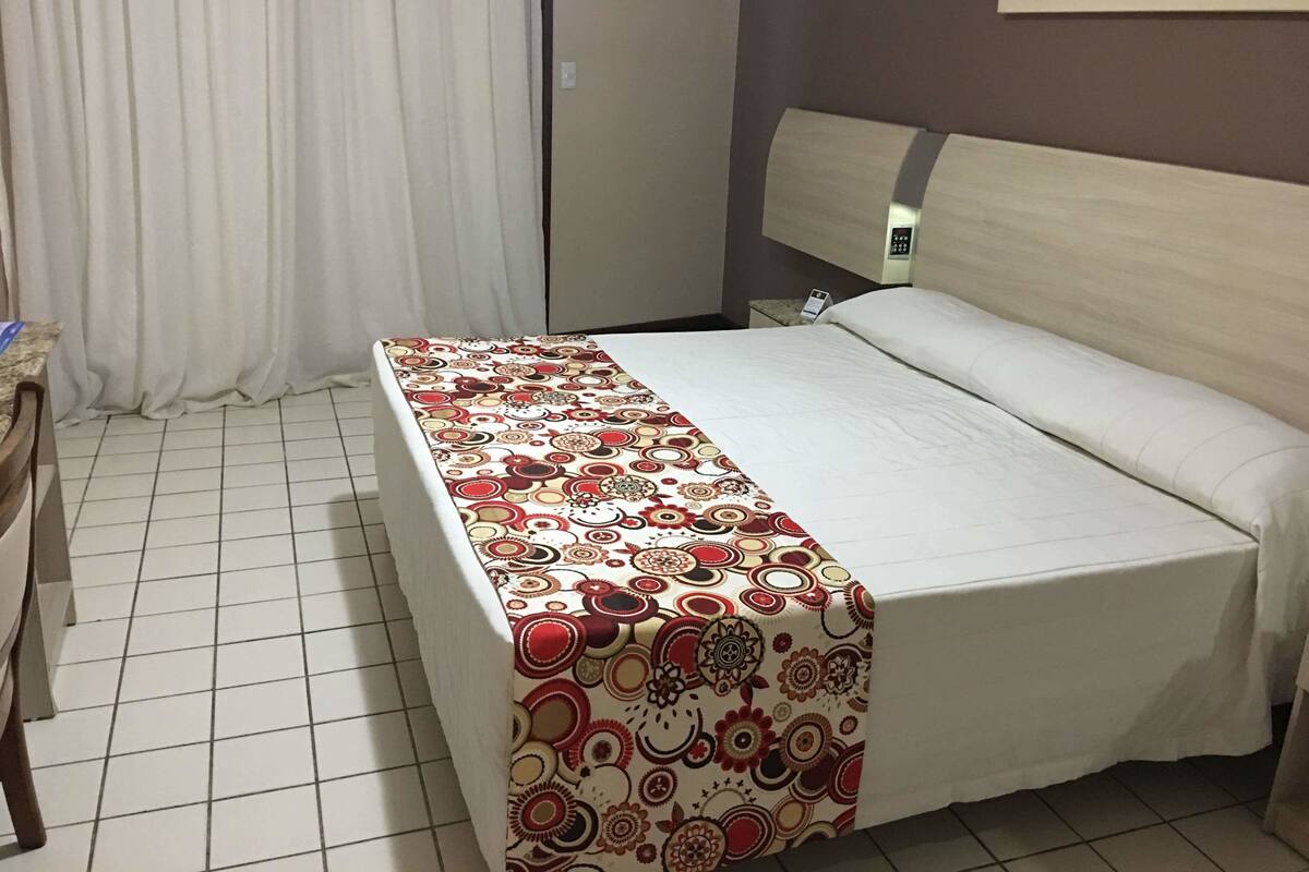 A hotel room with a bed, a desk, a chair, a curtain, and a painting on the wall.