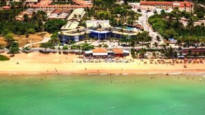A coastal resort with a sandy beach, a large building with a blue and white facade, and a palm tree-lined promenade. - Sueds Plaza (Porto Seguro)
