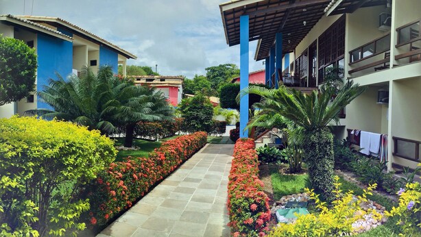 A courtyard with a paved walkway, surrounded by lush greenery and flowering plants. - Sueds Premium (Porto Seguro)