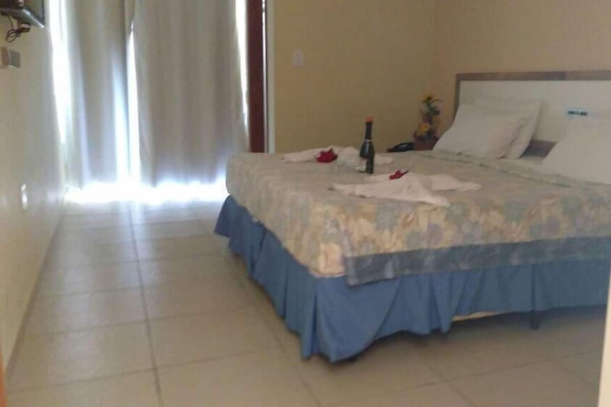 A hotel room with a bed, a bedside table, a window with curtains, and a tiled floor.