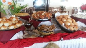 A buffet table with various food items and a glass of dark liquid, likely juice or wine, in a dining area with chairs and a vase of flowers. - Sueds Premium (Porto Seguro)