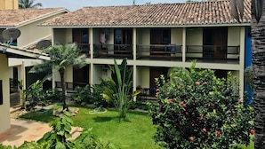 A two-story building with a tiled roof, multiple balconies, and a garden with various plants and a small tree. - Sueds Premium (Porto Seguro)
