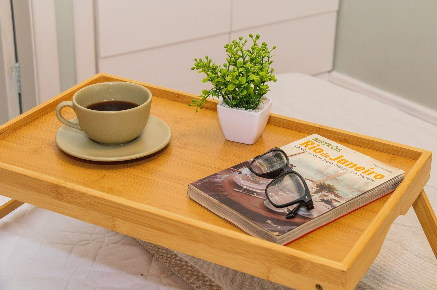 A wooden tray with a cup of coffee, a small potted plant, and a magazine with glasses on it.
