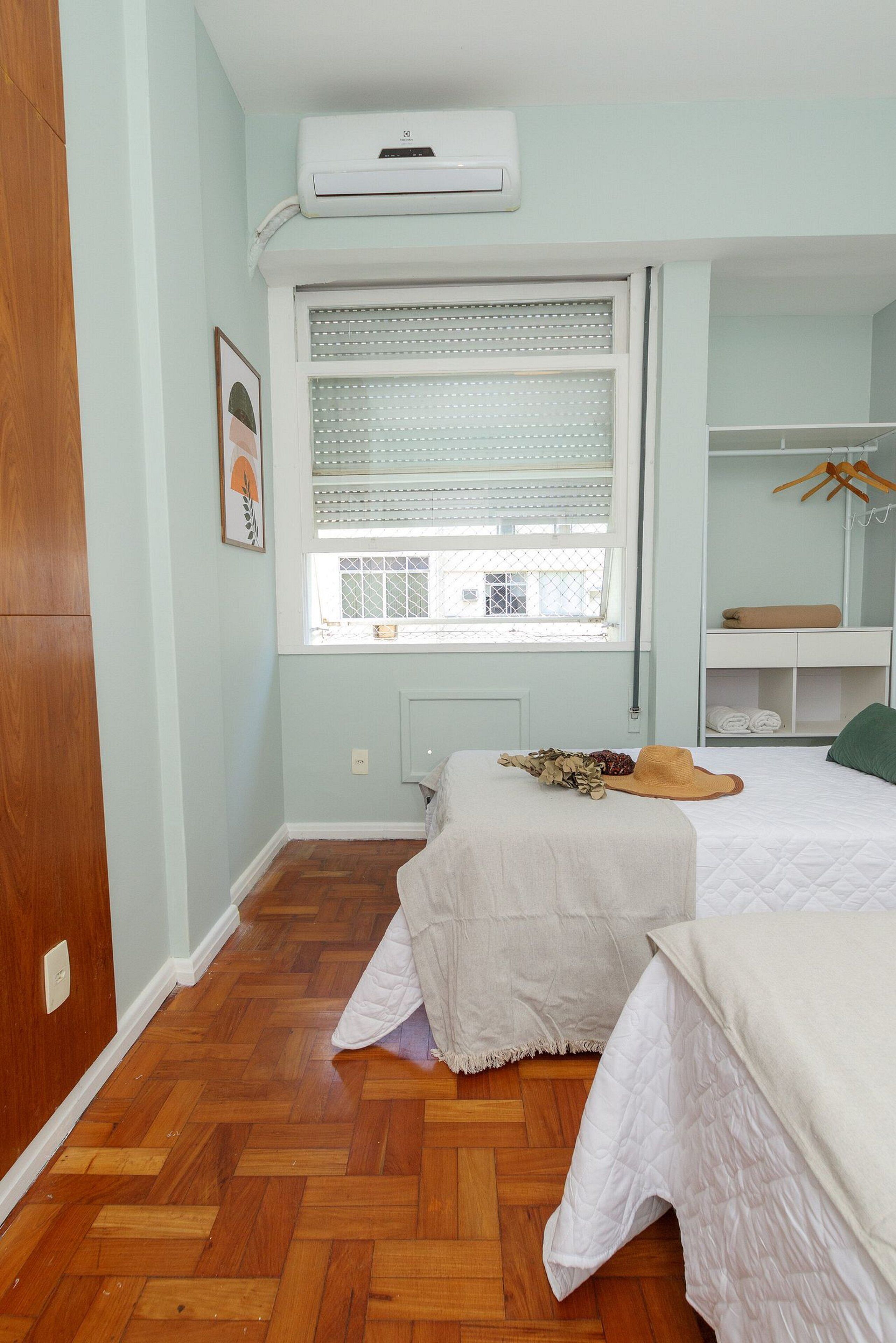 Een slaapkamer met raam, bed, nachtkastje, spiegel en een airconditioning.