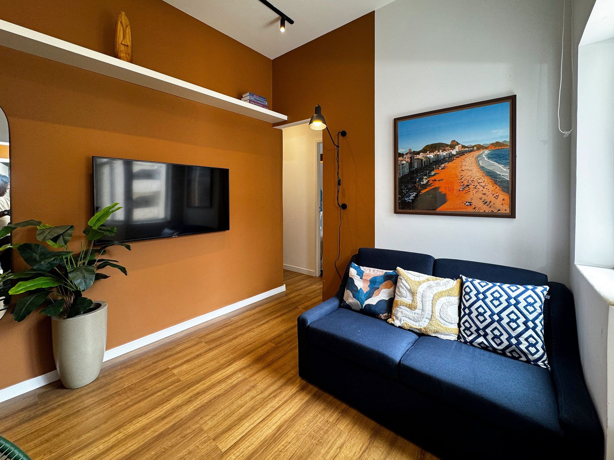 A modern living room with a flat-screen TV mounted on the wall, a blue sofa with patterned cushions, a wooden floor, and a painting of a beach scene.
