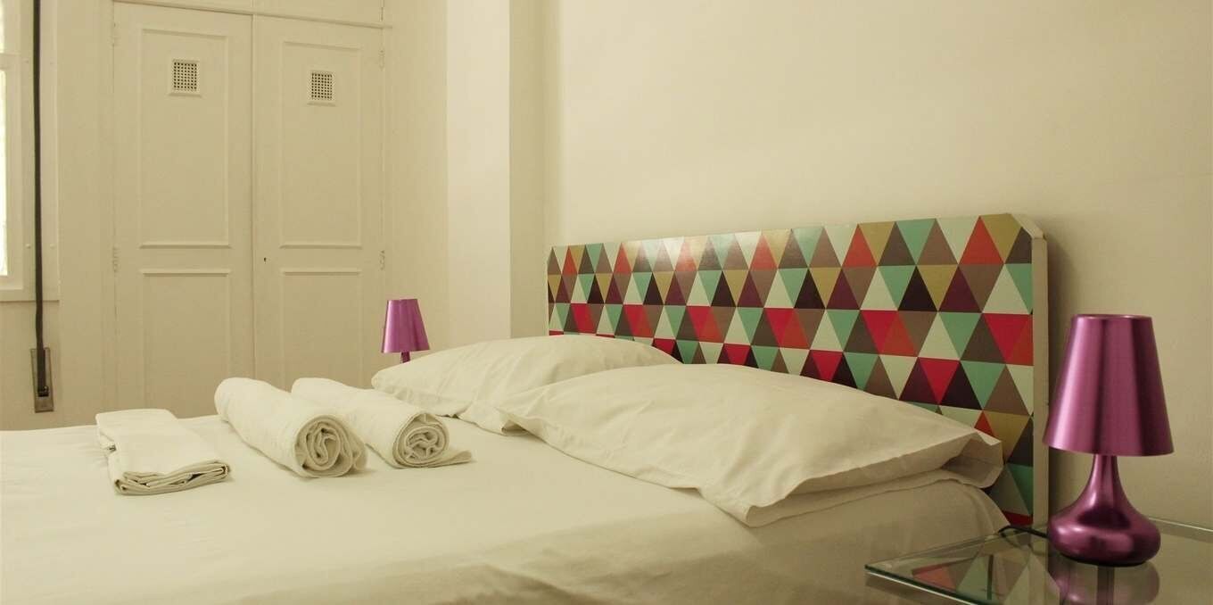 A hotel room with a bed, pillows, rolled towels, a bedside table with a lamp, and a colorful geometric-patterned headboard.