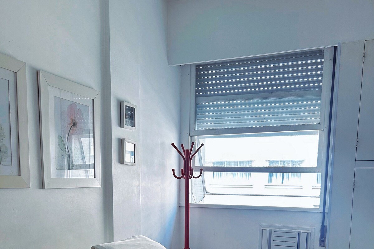 Un lit recouvert d’une couverture rouge, avec des oreillers blancs et des serviettes enroulées. Il y a une fenêtre avec des stores, un porte-manteau et des tableaux encadrés accrochés au mur.
