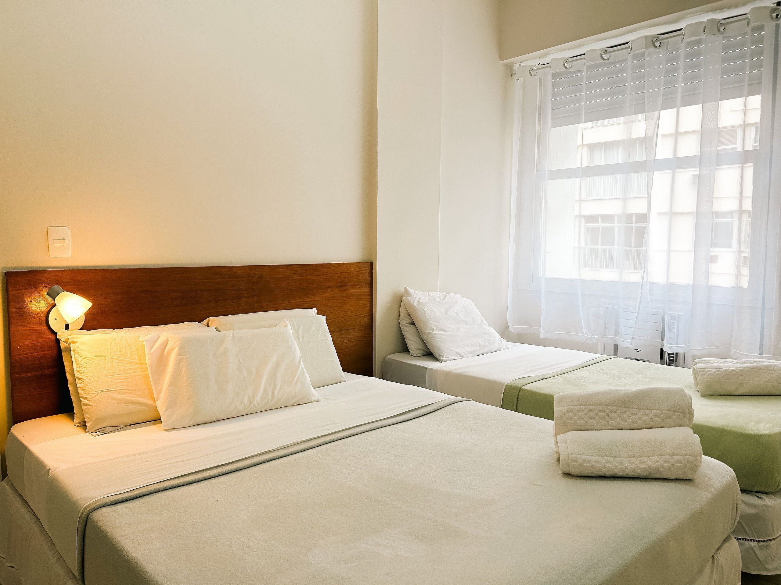 A hotel room with two beds, a wooden headboard, a bedside lamp, and a window with sheer curtains.