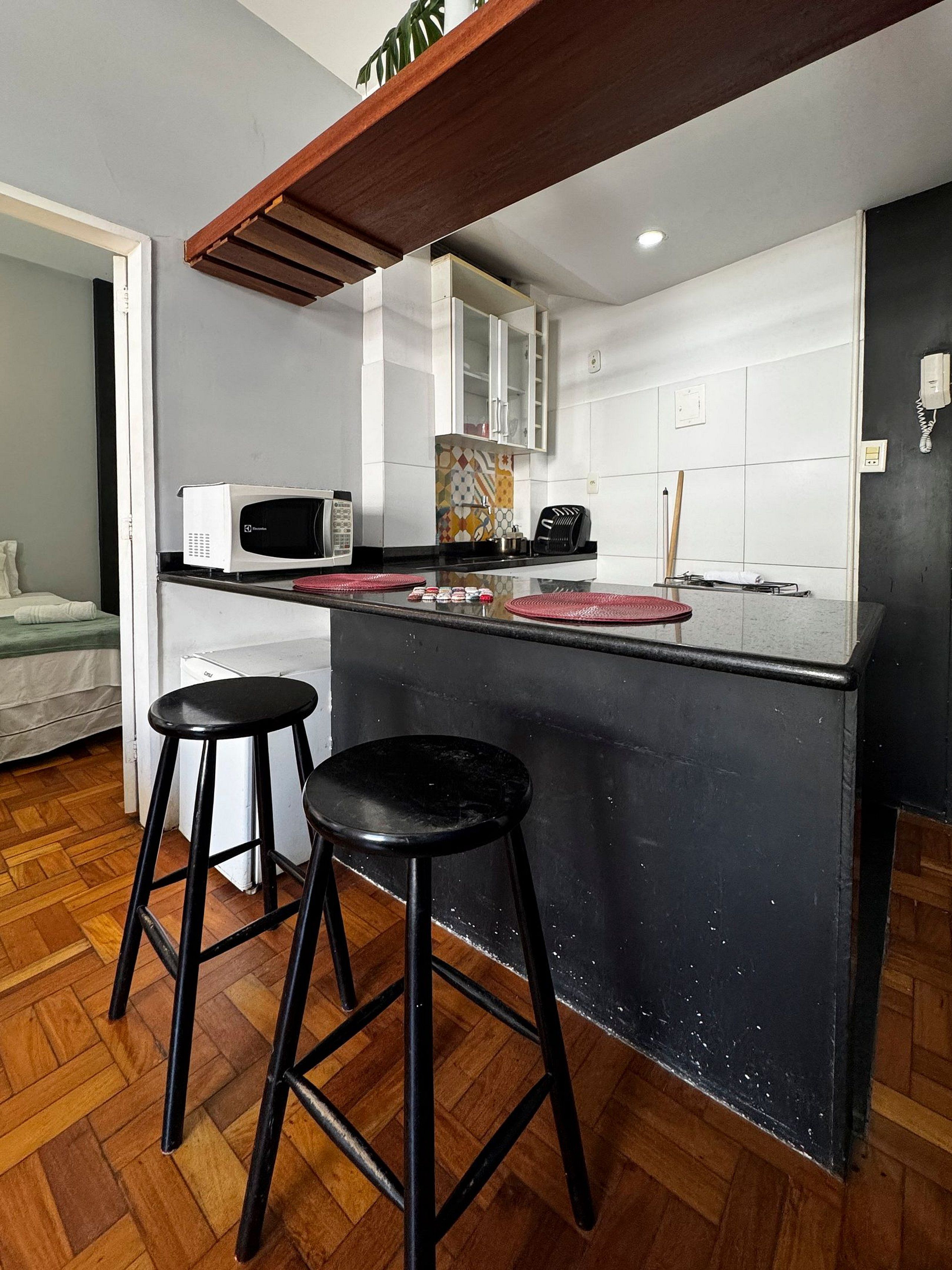 A compact kitchen with a black countertop, two stools, a microwave, and a cabinet.