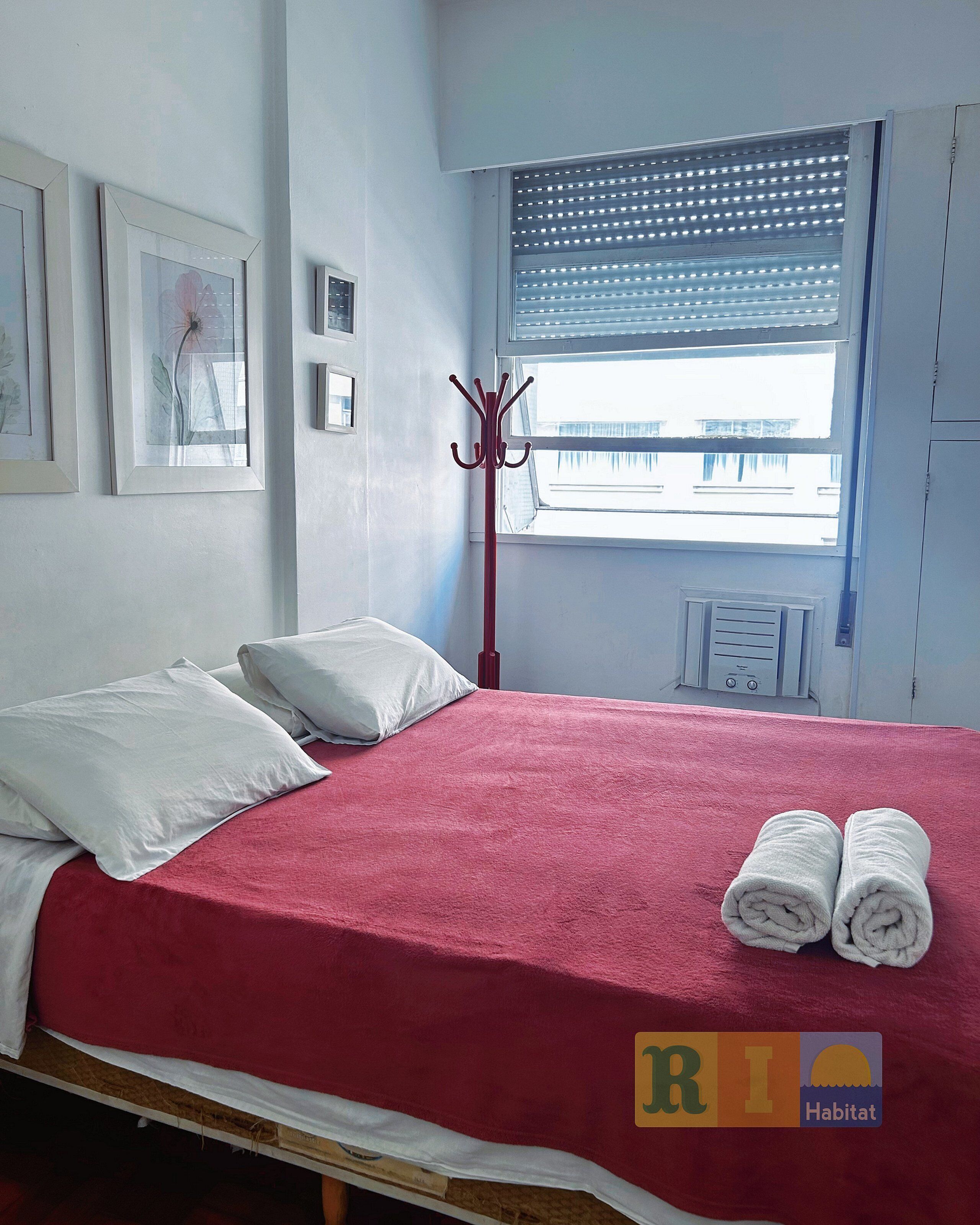A bed with a red cover, white pillows, and rolled towels. There is a window with blinds, a coat rack, and framed pictures on the wall.