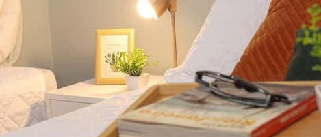 A neatly made bed with a white comforter, a bedside table with a lamp, a framed picture, and a small plant.