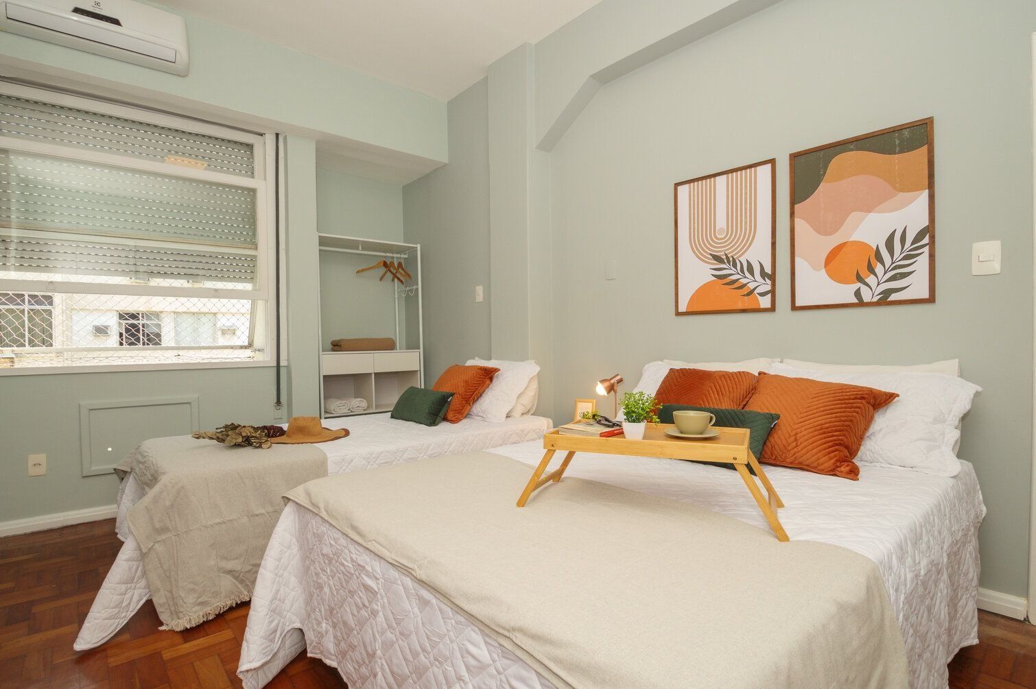 A bedroom with two beds, a window with blinds, a bedside table with a lamp, and artwork on the wall.